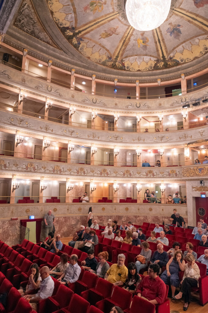 Blick in von der Seite in das Publikum im Theatro Imola. Im oberen Teil des Bildes ist die Decke und ein Kronleuchter, im mittleren Teil die Balkone des Theaters zu sehen